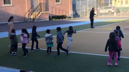 Children in a line follow a leader's movements on a colorful outdoor CommonPoint Challenge court.